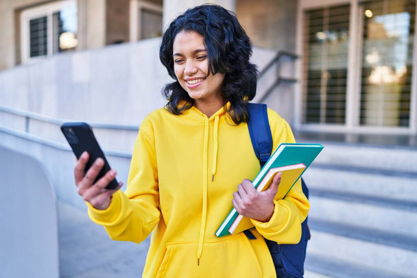 Young latin woman student holding books using smartphone at street - Photo, Image