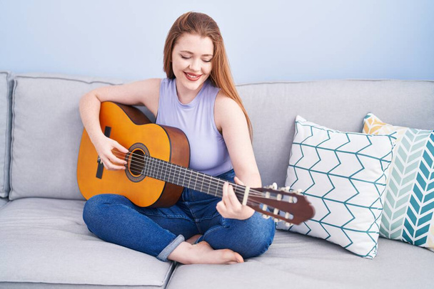 Young redhead woman playing classical guitar sitting on sofa at home - Fotó, kép