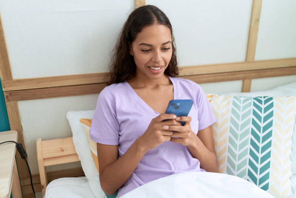 Young african american woman using smartphone sitting on bed at bedroom - Foto, immagini