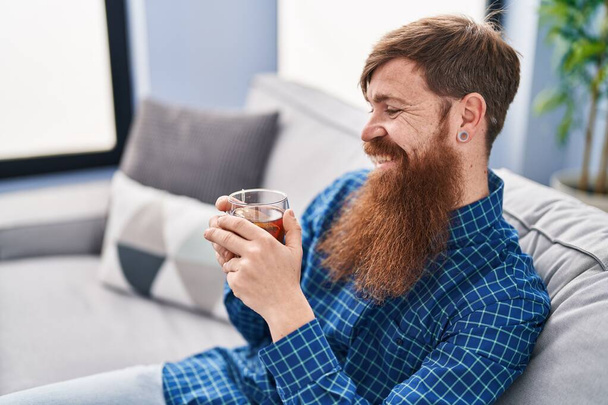 Νεαρός κοκκινομάλλης άνδρας πίνοντας τσάι κάθεται στον καναπέ στο σπίτι - Φωτογραφία, εικόνα