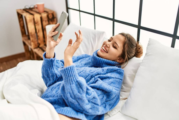 Νεαρή όμορφη ισπανόφωνη γυναίκα που χρησιμοποιεί smartphone ξαπλωμένη στο κρεβάτι στο υπνοδωμάτιο - Φωτογραφία, εικόνα