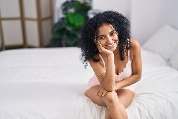 Jovem hispânica sorrindo confiante sentado na cama no quarto - Foto, Imagem