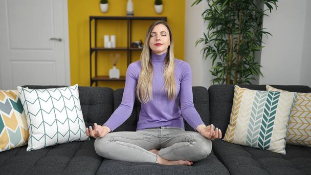Young blonde woman doing yoga exercise sitting on sofa at home - Valokuva, kuva