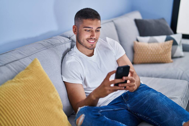 Jovem hispânico usando smartphone sentado no sofá em casa - Foto, Imagem