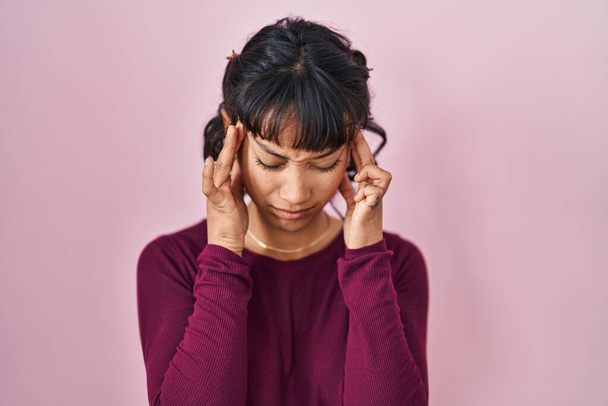 Jonge mooie vrouw staat over roze achtergrond met de hand op het hoofd voor pijn in het hoofd omdat stress. lijden aan migraine.  - Foto, afbeelding