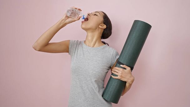 Young beautiful hispanic woman wearing sportswear holding yoga mat drinking water over isolated pink background - Foto, imagen