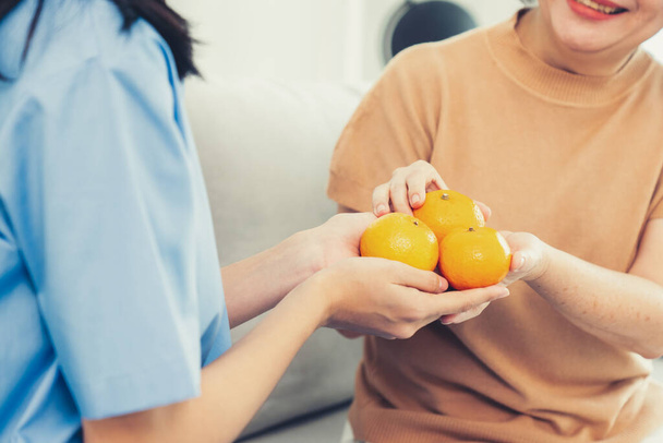 A young caregiver handing oranges to her contented senior patient at the living room. Senior care services, home visit by medical. - Valokuva, kuva