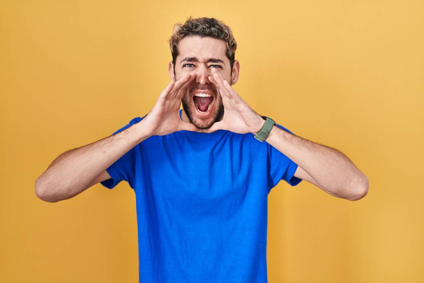 Hombre hispano con barba de pie sobre fondo amarillo gritando enojado en voz alta con las manos sobre la boca  - Foto, imagen
