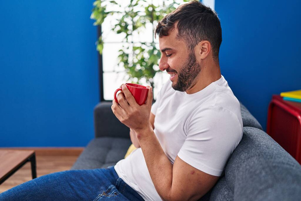 Junger hispanischer Mann trinkt Kaffee sitzend auf Sofa zu Hause - Foto, Bild