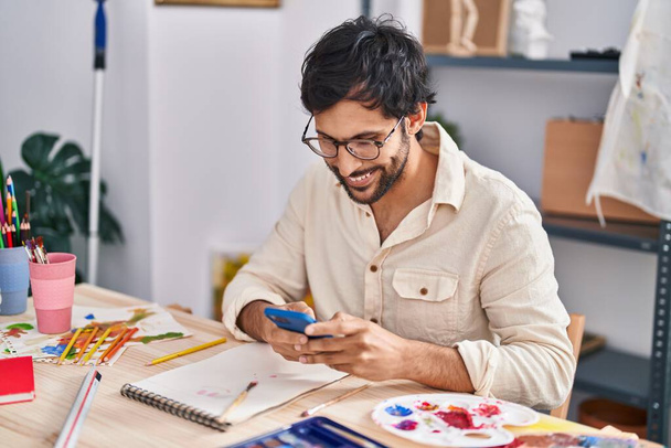młody Latynos człowiek artysta uśmiechnięty pewnie za pomocą smartfona w studio sztuki - Zdjęcie, obraz