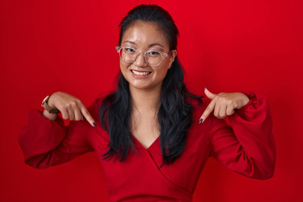 Aasialainen nuori nainen seisoo punaisella taustalla näyttäen luottavaiselta hymyillen kasvoilla, osoittaen itseään sormilla ylpeänä ja onnellisena.  - Valokuva, kuva