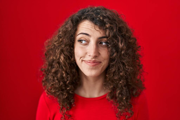 Mulher hispânica com cabelo encaracolado em pé sobre fundo vermelho sorrindo olhando para o lado e olhando para longe pensando.  - Foto, Imagem