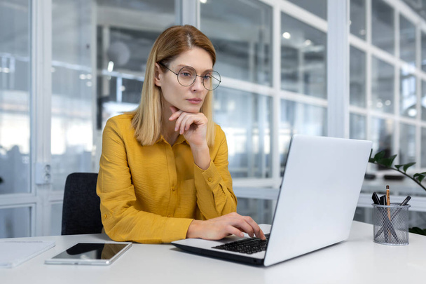 Portrait of a serious thinking businesswoman inside a bright office at work with a laptop, a woman in a yellow shirt and glasses works thoughtfully while sitting at a workplace - Foto, Imagen