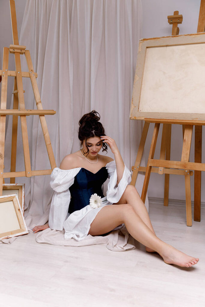 Ένα κορίτσι με μακριά σγουρά μαλλιά με μακιγιάζ σε λευκό φόρεμα με κορσέ κάθεται κοντά σε easels σε ένα στούντιο τέχνης. - Φωτογραφία, εικόνα