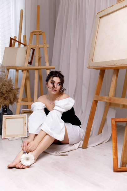 Девушка с длинными вьющимися волосами с макияжем в белом платье с корсетом сидит рядом с мольбертами в художественной студии. - Фото, изображение