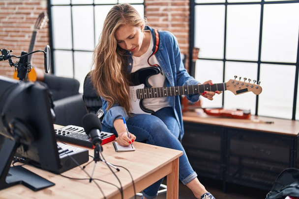 Young blonde woman musician composing song playing electrical guitar at music studio - Fotó, kép