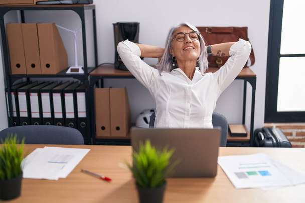 Moyen âge femme aux cheveux gris travailleur d'affaires en utilisant un ordinateur portable détendu avec les mains sur la tête au bureau - Photo, image