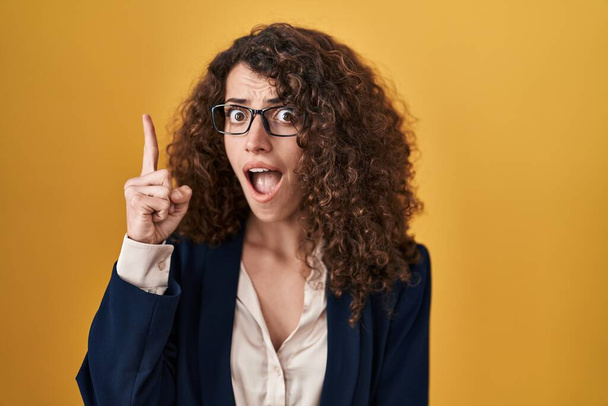 Hispanische Frau mit lockigem Haar steht über gelbem Hintergrund und zeigt mit erhobenem Zeigefinger auf erfolgreiche Idee. aufgeregt und glücklich. Nummer eins.  - Foto, Bild