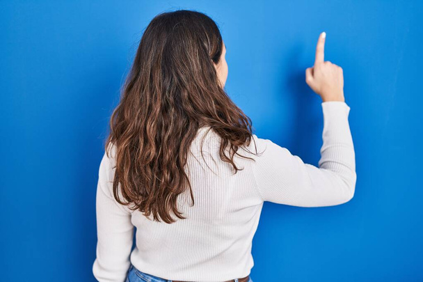 Junge hispanische Frau vor blauem Hintergrund posiert rückwärts und zeigt mit der Hand nach vorn  - Foto, Bild