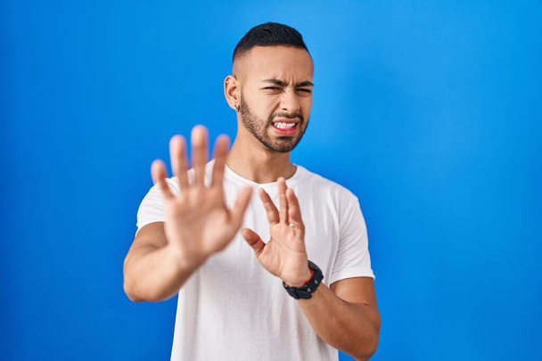 Young hispanic man standing over blue background disgusted expression, displeased and fearful doing disgust face because aversion reaction.  - Fotoğraf, Görsel