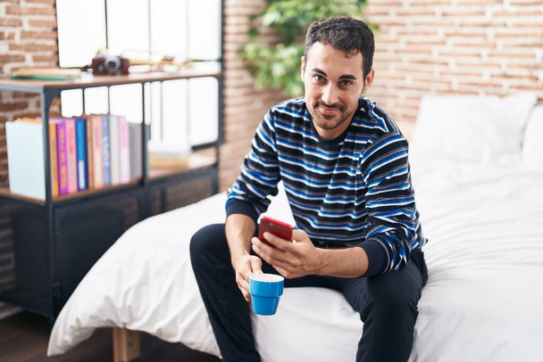 Young hispanic man using smartphone drinking coffee at bedroom - Foto, afbeelding