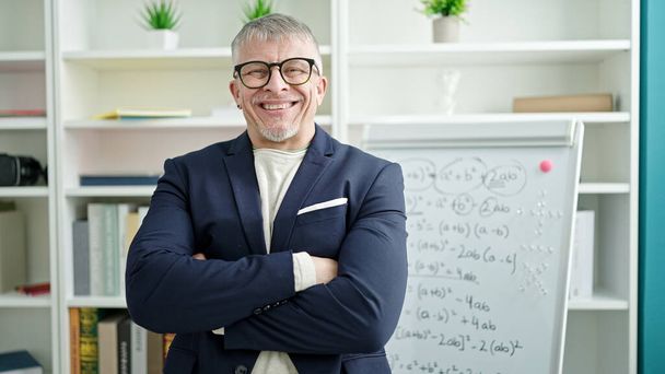 Μέση ηλικία γκριζομάλλης δάσκαλος άνθρωπος χαμογελά αυτοπεποίθηση στέκεται με τα χέρια σταυρωμένα χειρονομία από λευκό διοικητικό συμβούλιο στην τάξη του πανεπιστημίου - Φωτογραφία, εικόνα