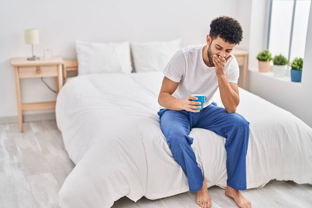 Young arab man tired drinking cup of coffee sitting on bed at bedroom - Photo, Image