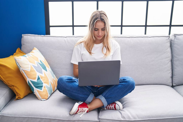 Молодая блондинка с ноутбуком сидит на диване дома - Фото, изображение