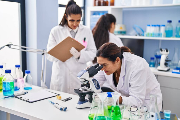 Three woman scientists using microscope write on checklist at laboratory - Foto, Imagem