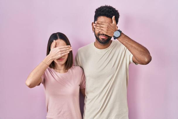 Jong Latijns-Amerikaans koppel samen over roze achtergrond bedekken ogen met de hand, kijken ernstig en verdrietig. gezichtsloosheid, verstoppertje en afwijzing  - Foto, afbeelding