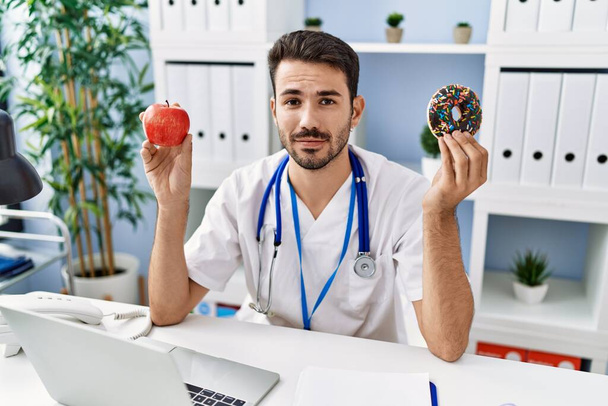 Young hispanic dietitian man holding doughnut and apple relaxed with serious expression on face. simple and natural looking at the camera.  - Φωτογραφία, εικόνα