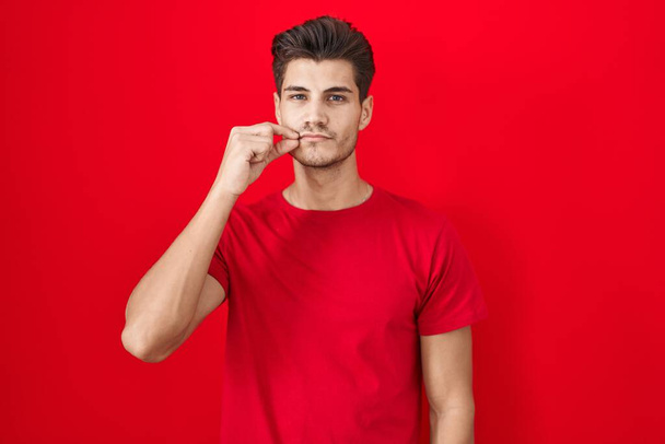 Fiatal spanyol férfi áll a piros háttér száj és az ajkak csukott, mint a zip ujjakkal. titkos és csendes, tabu beszél  - Fotó, kép