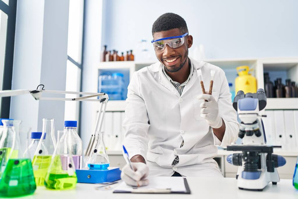 Young african american man wearing scientist uniform writing on clipboard holding test tubes at laboratory - Foto, immagini