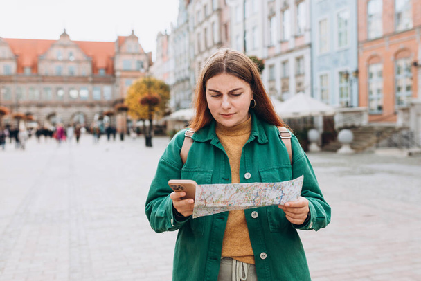 悲しい若い女性観光客が新しい都市を探索しています。赤い頭の女の子は古い町を歩いて、晴れた日に紙の地図とスマートフォンを保持. - 写真・画像