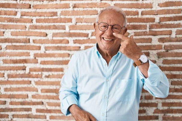 Hombre mayor con el pelo gris de pie sobre la pared de ladrillos señalando con el dedo de la mano a la cara y la nariz, sonriendo alegre. concepto de belleza  - Foto, Imagen