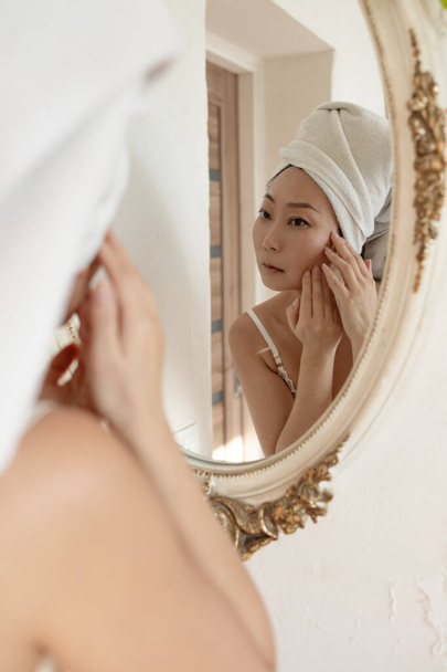 Portrait sad Asian middle aged woman looking in a mirror unhappy with wrinkles on her face. Skincare and aging concept. Human emotions face expression - Zdjęcie, obraz