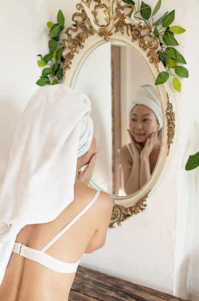 Happy confident young asian lady with towel on head looking at bathroom mirror, touching moisturized soft healthy sensitive skin, doing morning hydration, spa beauty treatment, face skin care concept - Foto, immagini