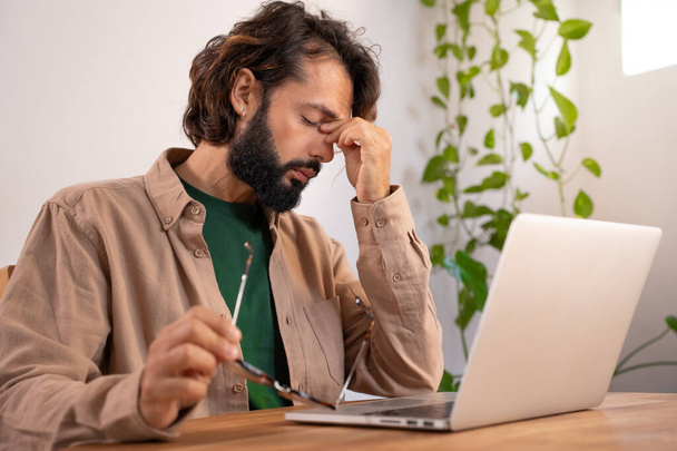 Stressed tired man suffering from headache in front of computer holding his glasses - Worried male entrepreneur thinking about problems and project deadline feeling exhausted. High quality photo - Φωτογραφία, εικόνα