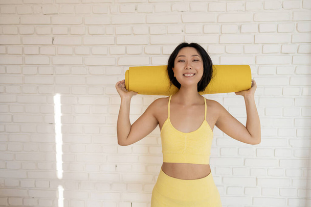 Happy attractive young asian woman holds in hands yellow yoga or fitness mat after working out at home in living room. Healthy life, keep fit concepts. - Фото, зображення