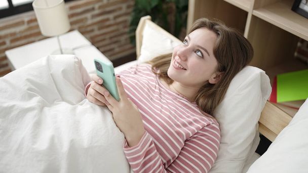 Junge blonde Frau mit Smartphone im Schlafzimmer liegend - Foto, Bild