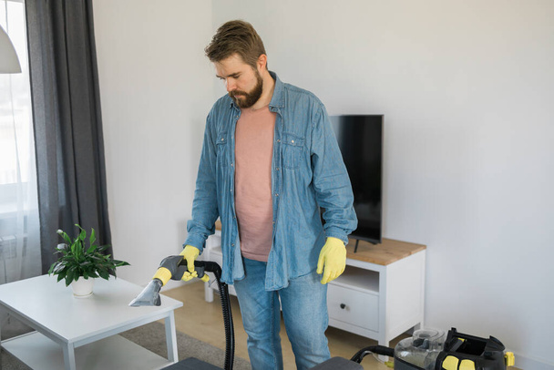 Muž držící moderní prací vysavač a čistící špinavou pohovku s profesionálně čistícím prostředkem. Profesionální jarní úklid doma - Fotografie, Obrázek