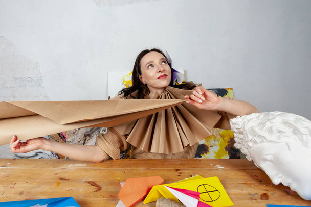 Portrait of a creative young woman sitting at a desk in her workshop, wearing a Tudor collar made of paper and holding huge paper plane. - Foto, Bild