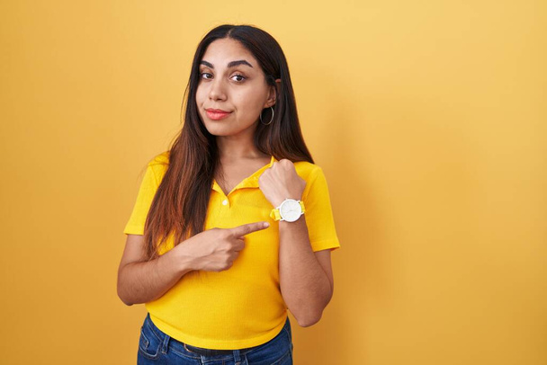 Young arab woman standing over yellow background in hurry pointing to watch time, impatience, looking at the camera with relaxed expression  - Photo, Image