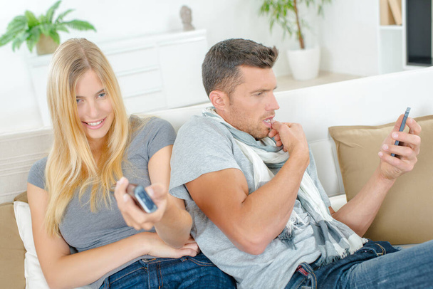 Couple on sofa, lady holding remote control, man looking at cellphone - Foto, Bild