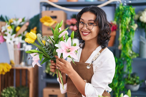 Young beautiful hispanic woman florist holding bouquet of flowers at flower shop - 写真・画像