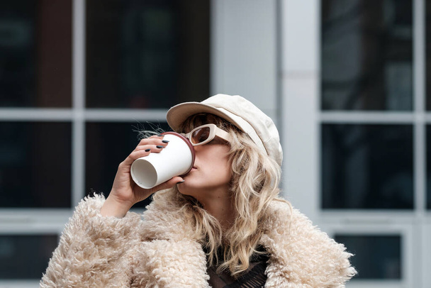 Modelo de mulher alternativa está bebendo café lá fora, em uma rua de Londres. É um dia frio de inverno e ela está usando um casaco pesado, boné e óculos de sol. - Foto, Imagem