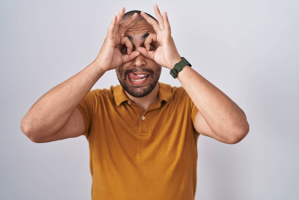 Hombre hispano con barba de pie sobre fondo blanco haciendo buen gesto como binoculares sacando la lengua, ojos mirando a través de los dedos. expresión loca.  - Foto, imagen