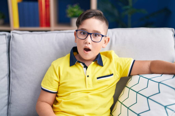Young hispanic kid sitting on the sofa at home scared and amazed with open mouth for surprise, disbelief face  - Zdjęcie, obraz