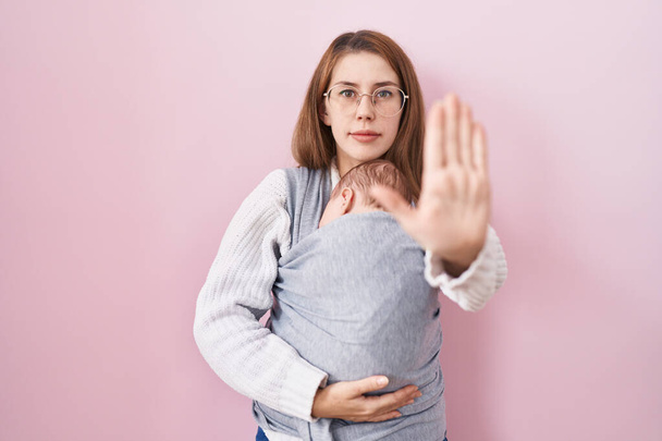 Fiatal kaukázusi nő kezében és terhes baba egy parittya nyitott kézzel csinál stop jel komoly és magabiztos kifejezés, védelmi gesztus  - Fotó, kép