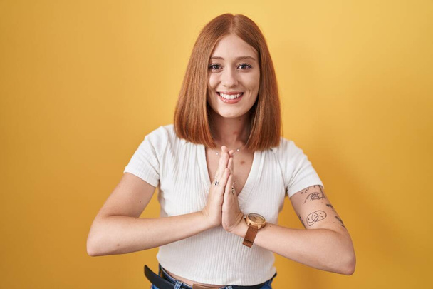 Young redhead woman standing over yellow background praying with hands together asking for forgiveness smiling confident.  - Photo, Image